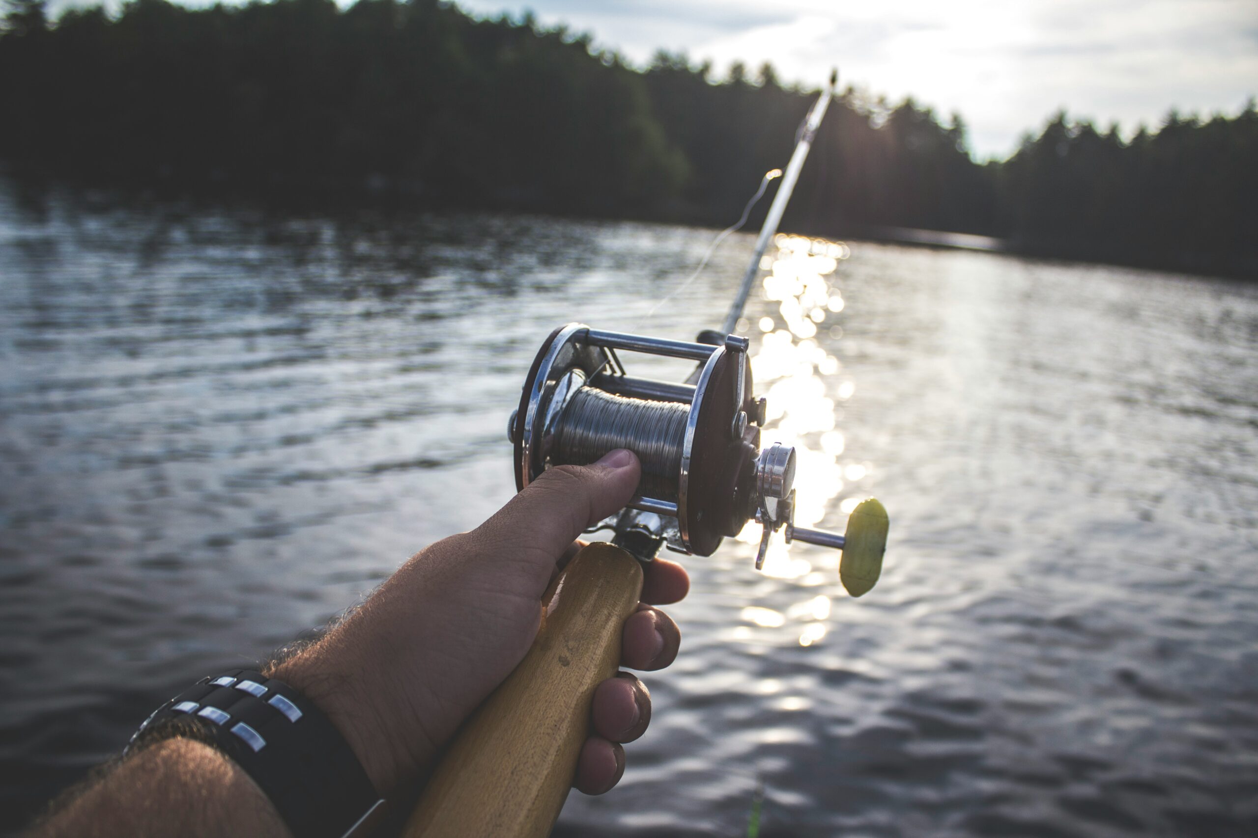 A close-up view of a person holding a fishing rod over a calm lake, with sunlight reflecting on the water.
