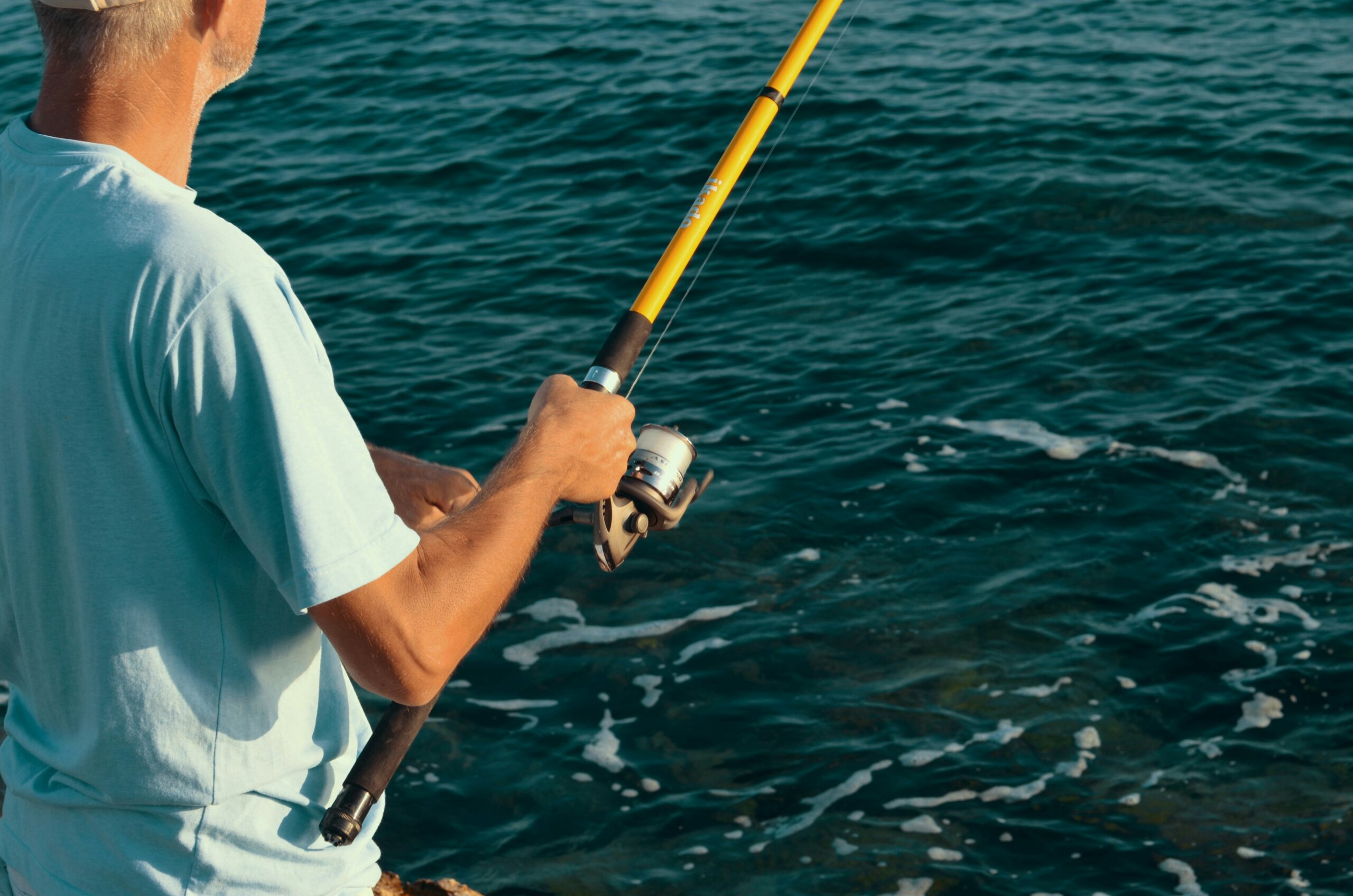 Man in a light blue t-shirt holding a yellow fishing rod while fishing near the shore on a calm sea.