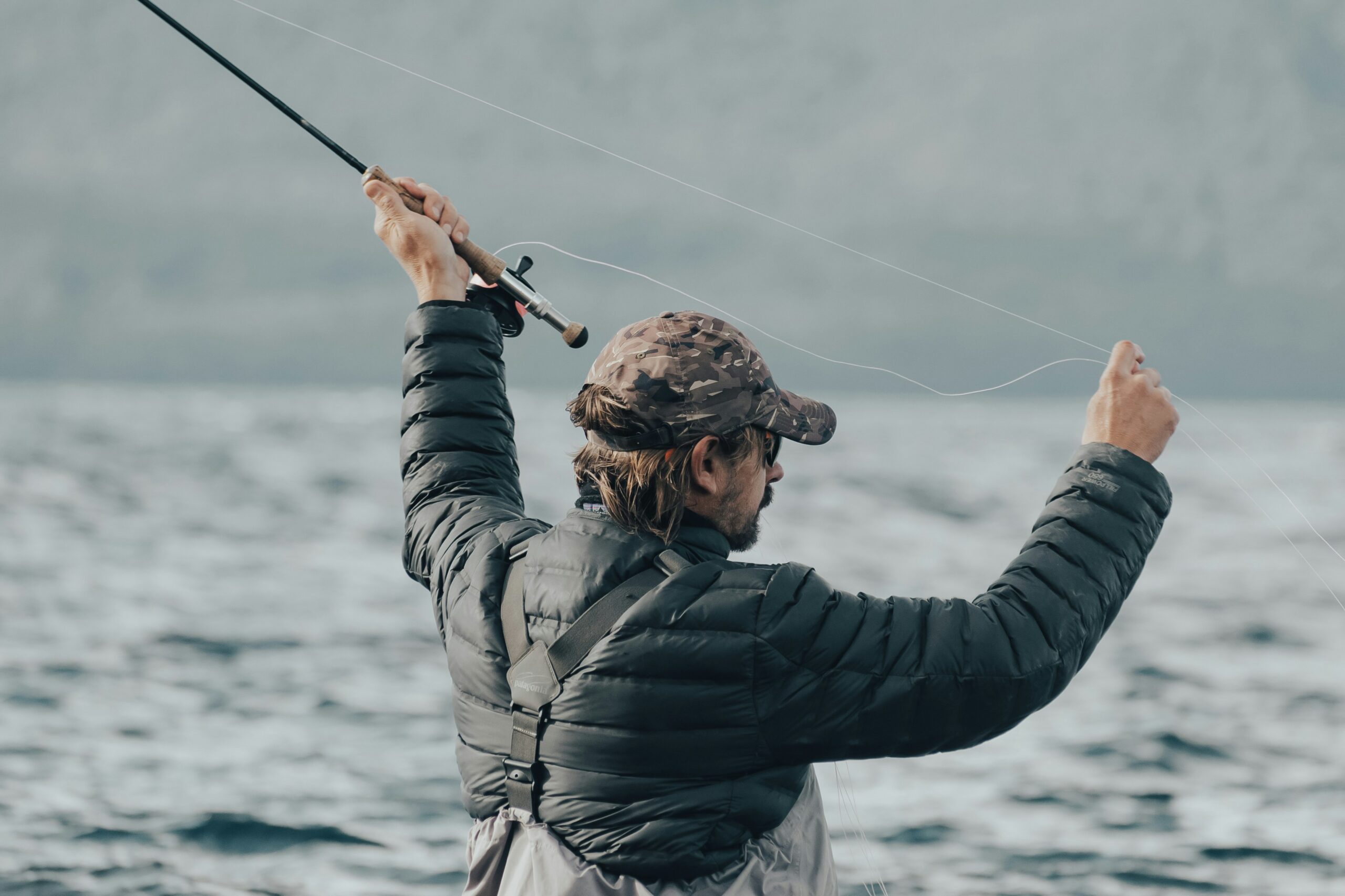 A person in a camouflage cap and black jacket fly fishing in open water with mountains in the background.

